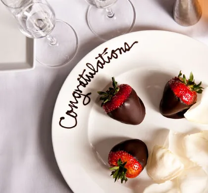 Three chocolate covered strawberries with white rose petals on a perfectly decorated white plate. Also to include a written congratulations in chocolate on the top of the plate.