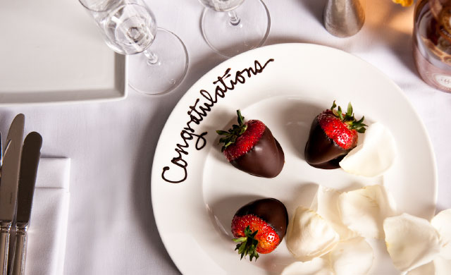 Three chocolate covered strawberries with white rose petals on a perfectly decorated white plate. Also to include a written congratulations in chocolate on the top of the plate.