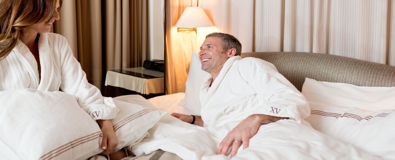 A couple both in our fifteen beacon robes laying in bed cozening up with our Frette linens.