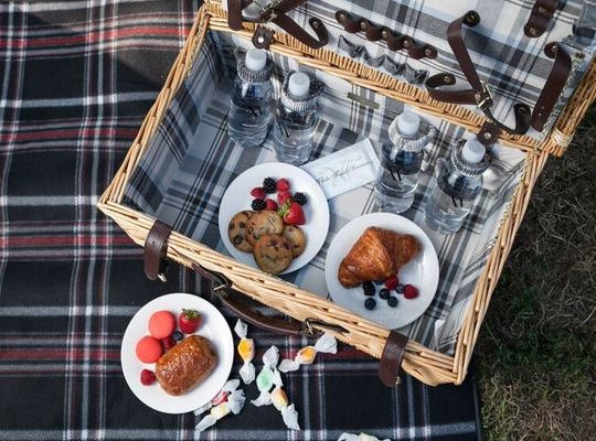 Open picnic basket with four bottle waters, chocolate chip cookies and a break croissant showcased in the inside of the basket.