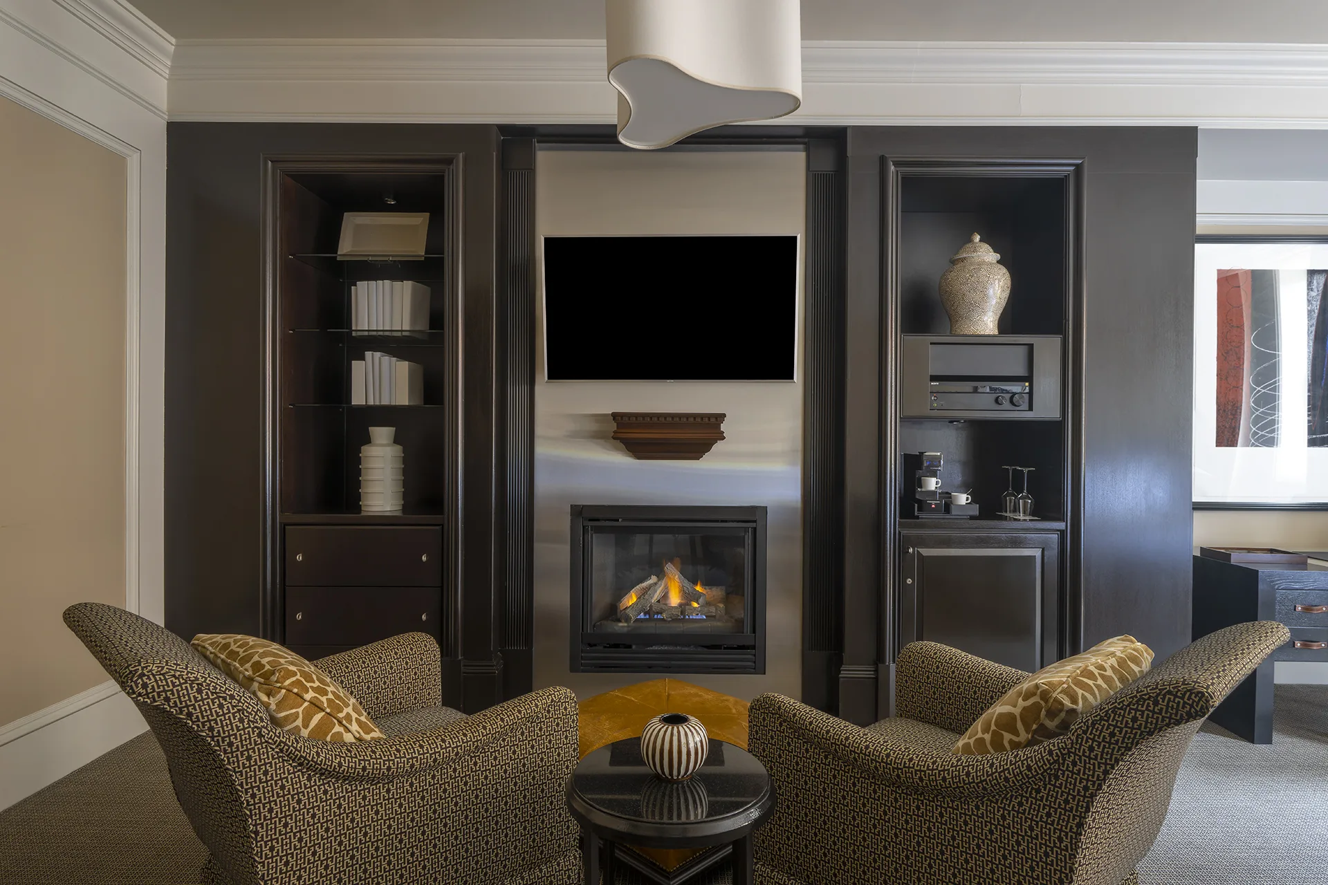 Studio room sitting area with two upholstered chairs, end table, dark wood accents, fireplace, and wall-mounted television.