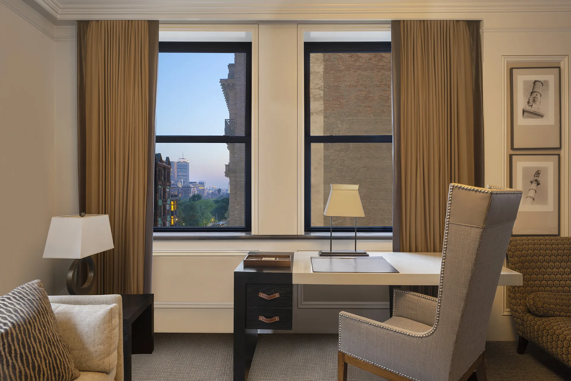 In-room workstation with a high-backed chair, desk, lamps, sofa and chaise lounge. A window overlooks boston behind the desk.