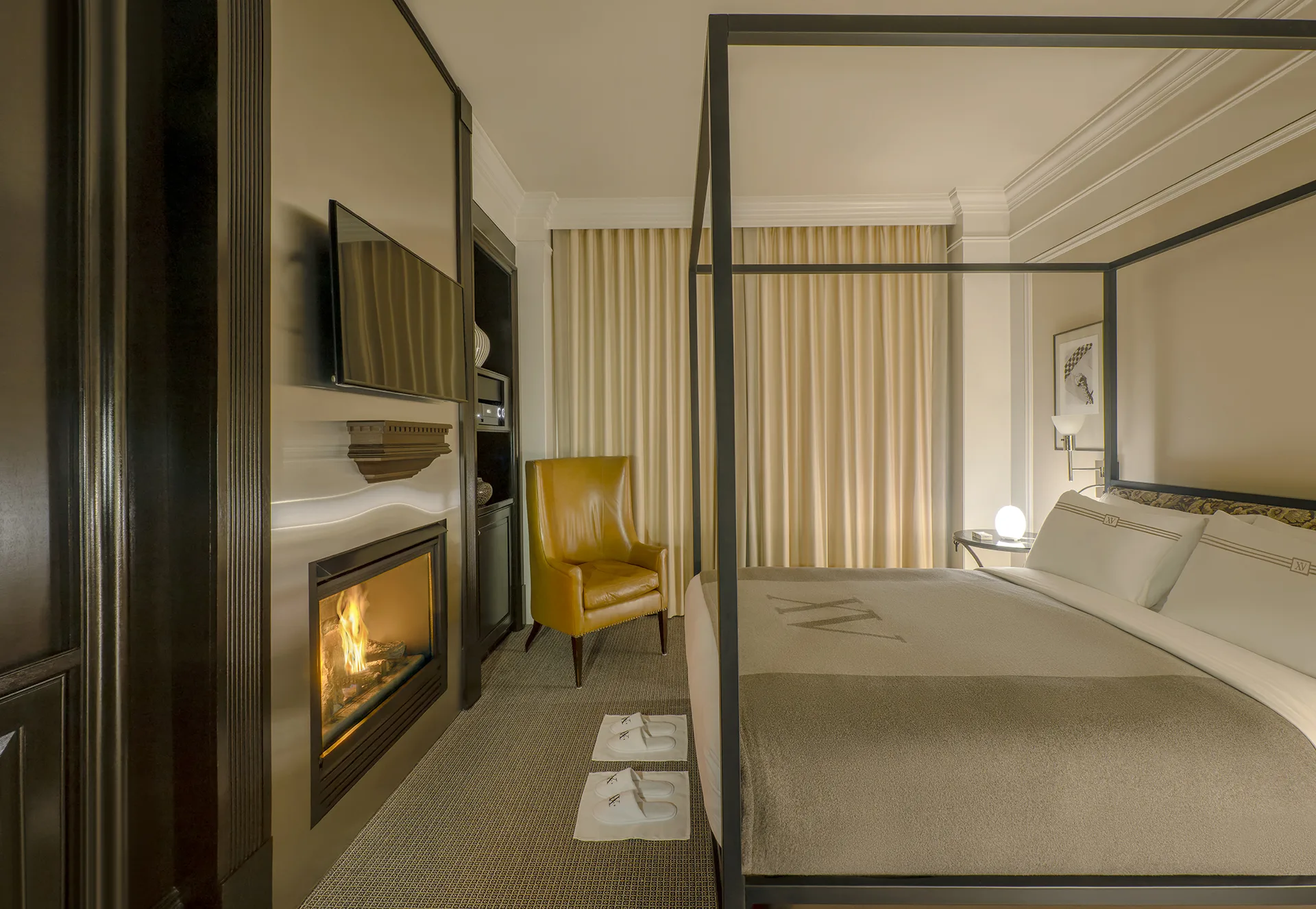 Guestroom at XV Beacon with monogrammed bedding, fireplace, chair, wall-mount TV and two pairs of guest slippers.