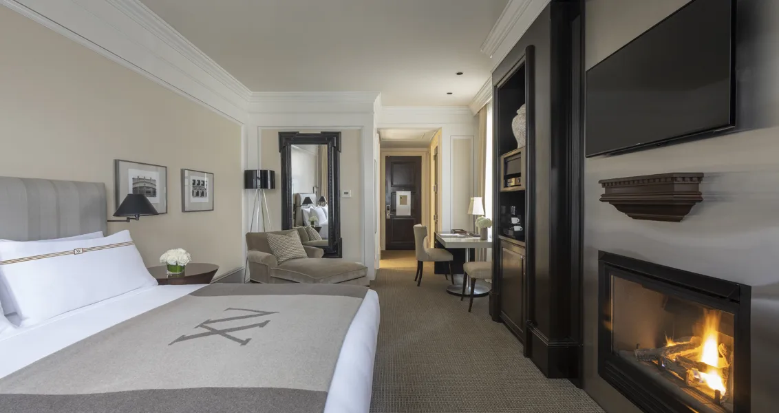 Interior view of a King room with monogrammed bedding, chaise lounge, fireplace, workstation, and wall-mounted television.