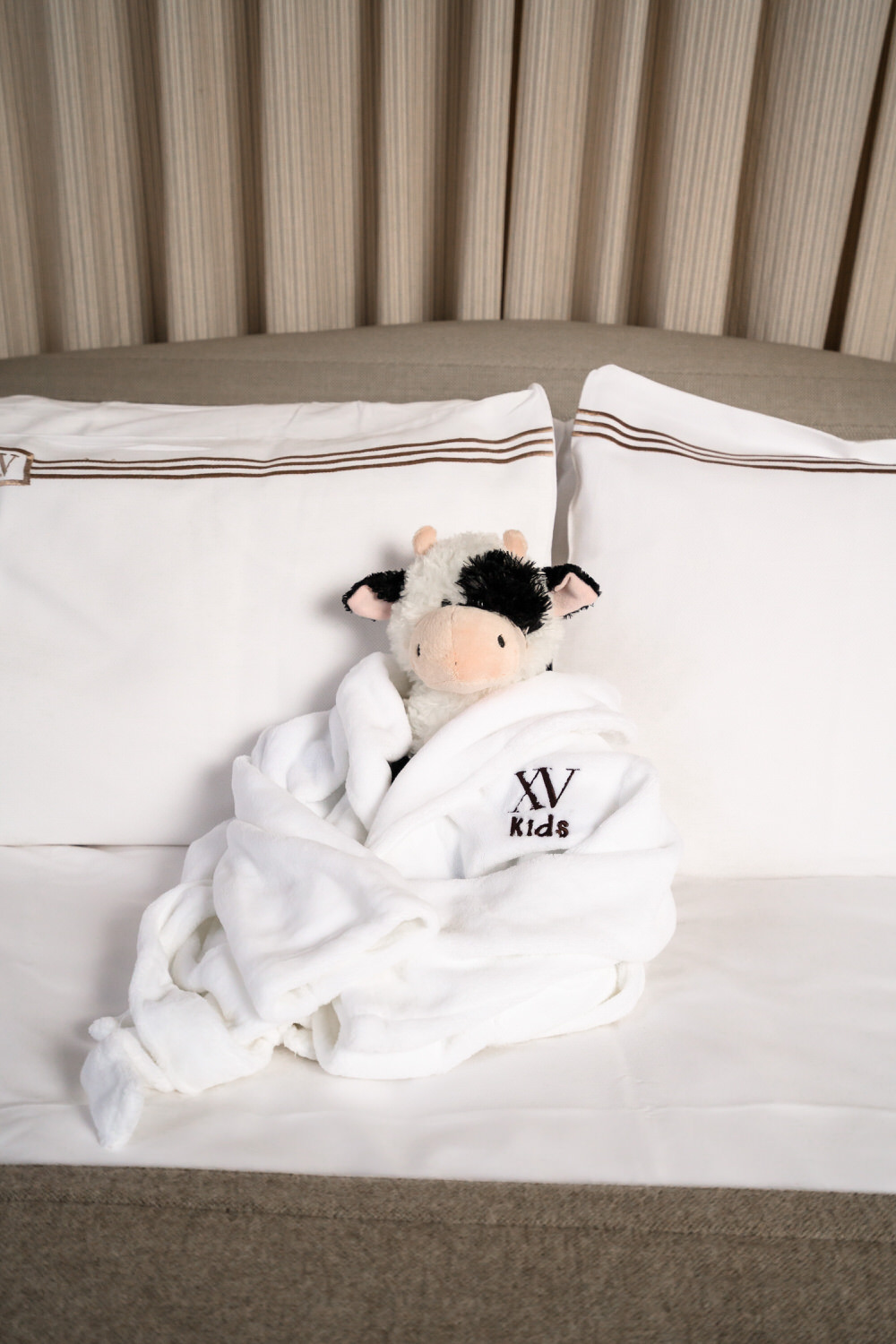 Stuffed animal cow dressed in an XV Beacon kids robe and sitting on the bed