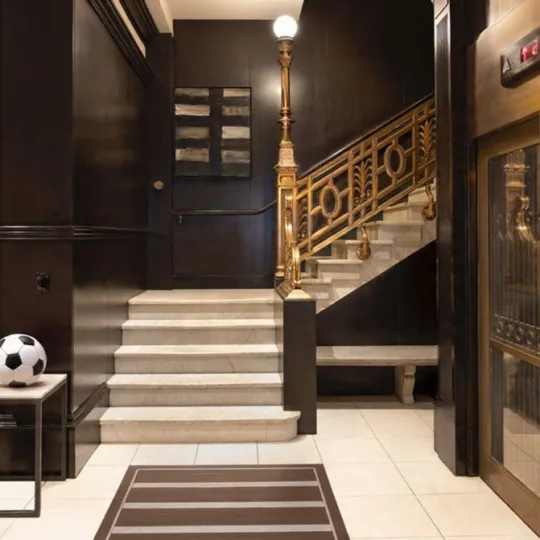 The lobby at XV Beacon with dark woods, an ornate staircase, glass elevator door, accent décor and light tile floor.